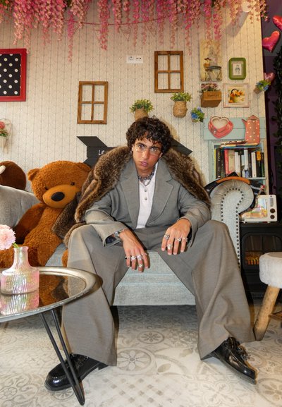 Traje gris de gran tamaño con un cuello de piel, superpuesto a una blusa gris. Los accesorios incluyen varios anillos y gafas. Sentado en una silla tapizada de color azul claro.