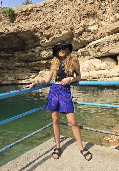 Sombrero de cubo negro, top negro sin mangas, pantalones cortos morados con rayas, sandalias negras. De pie en un muelle de piedras cerca de agua verde y acantilados.