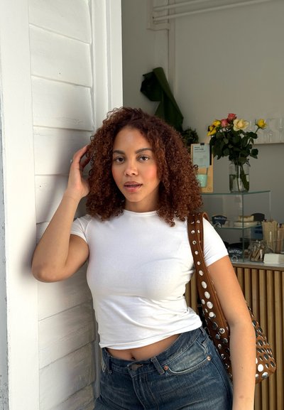 Camiseta blanca ajustada, jeans azules de cintura alta, cabello rizado. Bolso marrón perforado con acentos metálicos. Entorno interior con plantas.