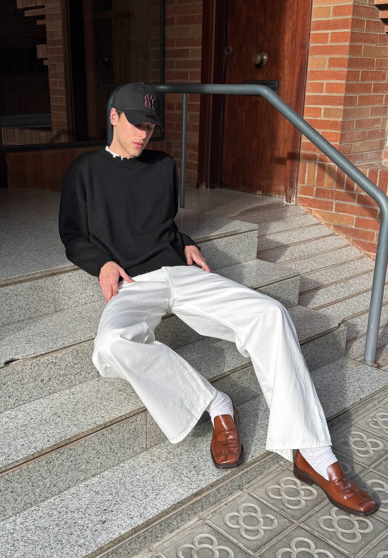 Black knitted jumper, white wide-leg jeans, brown leather shoes, sitting on stone steps with a grey railing in a sunlit area.