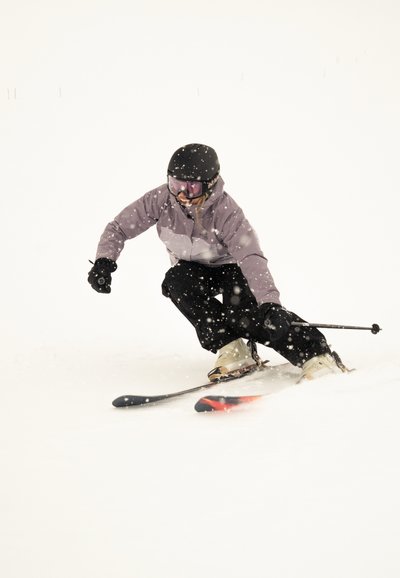 Esquiador con casco negro y gafas, chaqueta morada y pantalones negros, esquiando entre la nieve en condiciones nevadas.