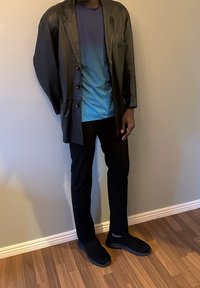 Manteau en cuir noir avec une poche unique, porté sur une chemise dégradée bleue. Pantalon noir ajusté et chaussures noires, debout sur un sol en bois.