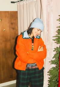 Personne souriante portant un bonnet bleu et une veste orange, les bras croisés, debout à côté de rideaux roses et d'un sapin vert partiel.