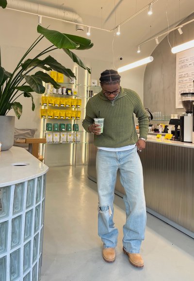Suéter verde olivo, jeans desgastados azul claro y calzado marrón; sosteniendo una bebida verde en una cafetería moderna con plantas y productos exhibidos.