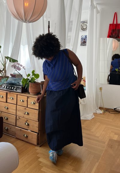 Camisa de rayas azul y blanca con un diseño drapeado, falda larga azul marino y zapatillas azules; cómoda de madera en una habitación luminosa y ventilada.
