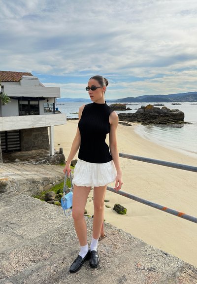 Top negro de canalé sin mangas, falda mini blanca con capas, mocasines negros gruesos, calcetines blancos y un pequeño bolso azul. Playa y rocas de fondo.