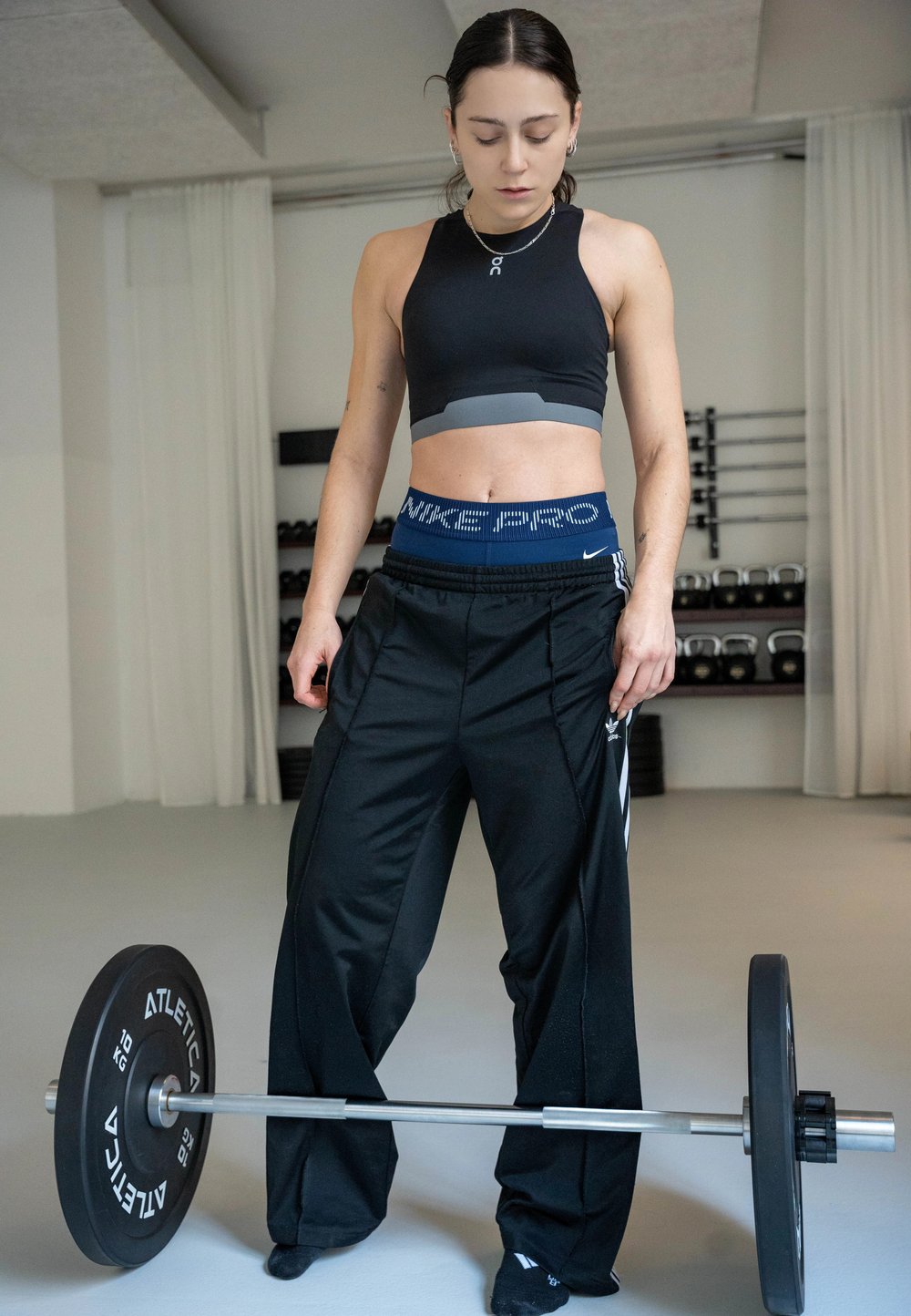 Donna in abbigliamento sportivo in piedi in palestra che guarda verso il basso un bilanciere con pesi su un pavimento grigio chiaro, con scaffali con kettlebell sullo sfondo.