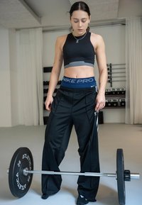 Donna in abbigliamento sportivo in piedi in palestra che guarda verso il basso un bilanciere con pesi su un pavimento grigio chiaro, con scaffali con kettlebell sullo sfondo.