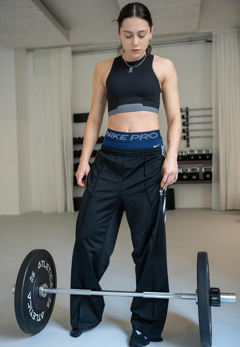 Γυναίκα σε αθλητική ενδυμασία που στέκεται σε γυμναστήριο και κοιτάζει προς τα κάτω μια μπάρα με βάρη, πάνω σε ανοιχτό γκρι πάτωμα, με ράφια που έχουν αλτήρες πίσω της.