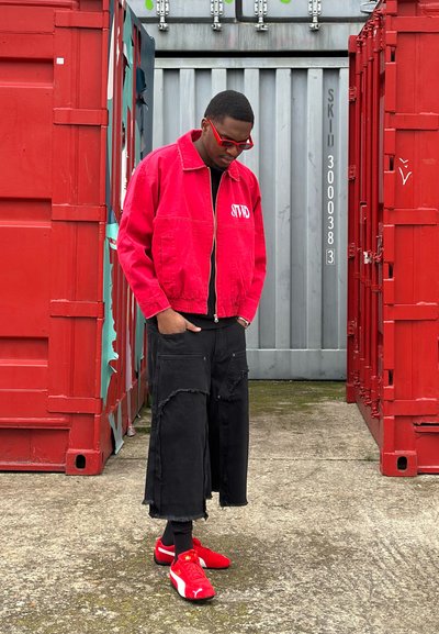 Chaqueta roja con cremallera y letras "STWD", pantalones negros de gran tamaño con bordes deshilachados, y zapatillas rojas, todo frente a contenedores de envío rojos.