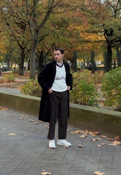 Suéter blanco con ribetes oscuros, pantalones burdeos, abrigo negro largo y zapatillas blancas, en un fondo de parque otoñal.