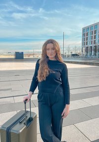 Hard-shell olivengrønn koffert med ribbet tekstur og svart håndtak. En stående kvinne i svart topp og bukser på et asfaltert område med blå himmel.