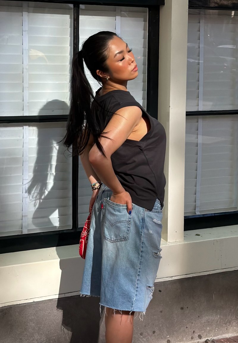 Femme avec une longue queue de cheval noire portant un haut noir sans manches et une jupe en jean déchirée, debout en pleine lumière près d'une fenêtre avec des stores blancs.