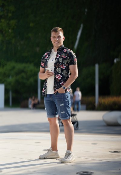 Joven masculino que lleva una camisa negra con flores sobre una camiseta blanca, pantalones cortos de mezclilla azul desgastados y zapatillas crema, sosteniendo un teléfono inteligente.