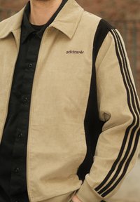 Veste en velours côtelé beige, avec un panneau noir et trois bandes sur les manches. Logo Adidas violet sur le côté gauche de la poitrine.