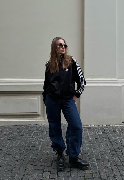 Chaqueta deportiva negra con rayas blancas, combinada con jeans azules sueltos y botas negras. El modelo tiene el cabello largo y lleva gafas de sol.