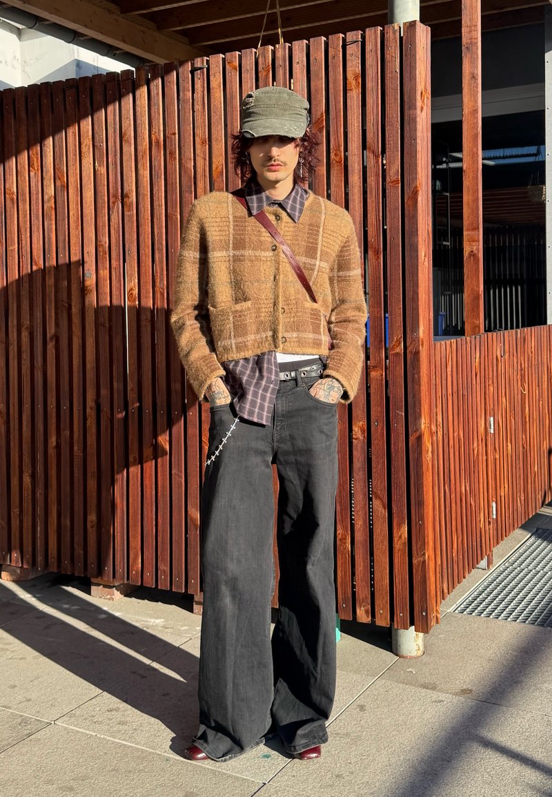 Persona che indossa un cardigan corto a quadri marrone, una camicia a quadri viola, pantaloni neri a gamba larga, un berretto verde e scarpe scure, in piedi contro una recinzione in legno.