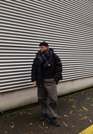 Chaqueta negra acolchada con un gran cuello, gorro de punto negro, pantalones cargo grises y zapatillas negras. El fondo presenta paredes acanaladas.