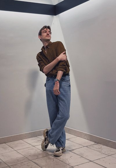 Camisa marrón de botones, jeans sueltos de un azul claro y zapatillas negras y blancas. Textura suave con un corte casual. De pie contra una pared neutra.