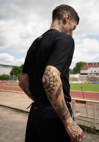 Camisola atlética preta com mangas curtas, apresentando um padrão texturizado; tatuagens proeminentes no braço direito incluem um padrão de teia de aranha e motivos da natureza.