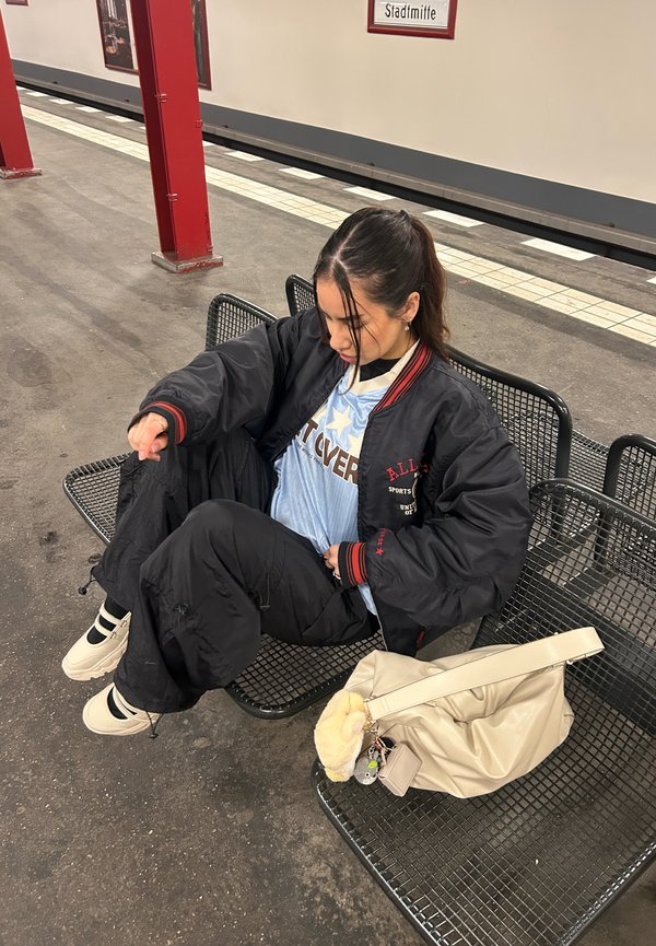 Jeune femme en veste et pantalon noirs assise sur un banc en métal sur le quai de la station de métro Stadtmitte, avec un sac beige à côté d'elle.