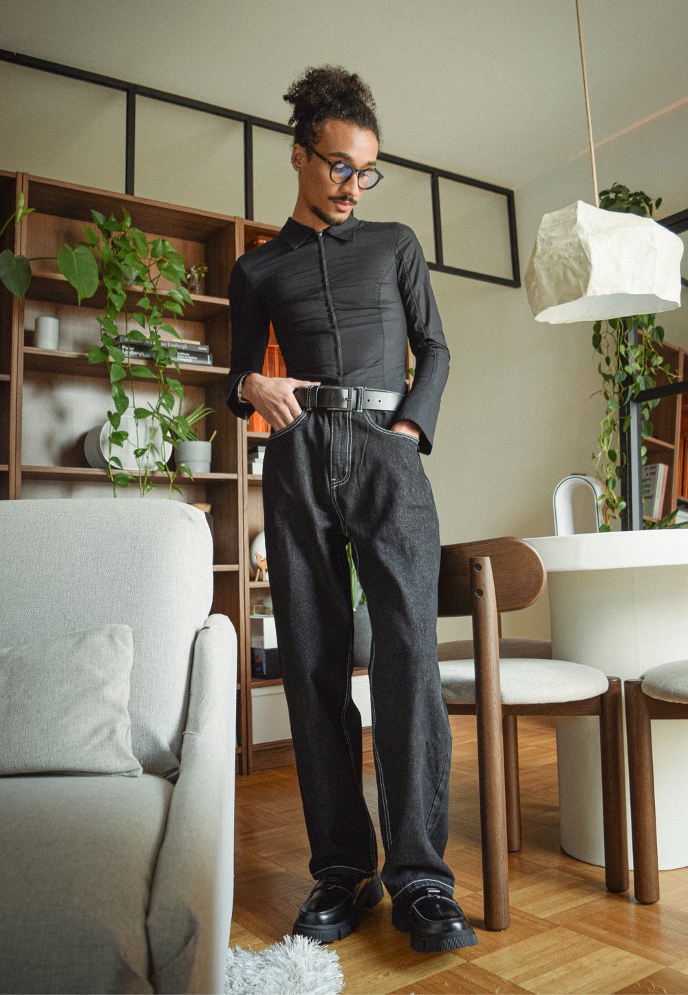 Zwart top met lange mouwen en een geknoopte kraag, donkerblauwe spijkerbroek met hoge taille en witte stiksels, en zwarte schoenen op een houten vloer.