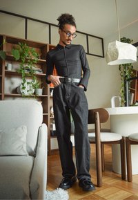 Zwart top met lange mouwen en een geknoopte kraag, donkerblauwe spijkerbroek met hoge taille en witte stiksels, en zwarte schoenen op een houten vloer.