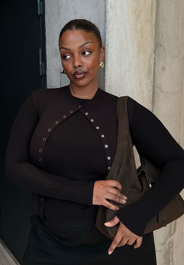 Femme aux cheveux tirés en arrière, portant un haut noir avec des œillets en métal, tenant un sac à bandoulière marron foncé contre un mur texturé en arrière-plan.