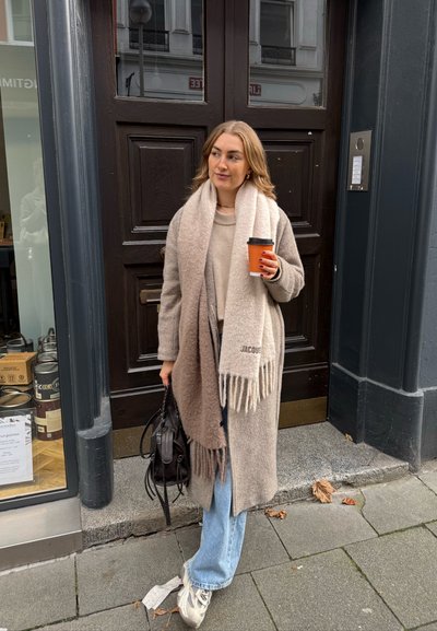 Mujer con abrigo beige y bufanda sostiene una taza de café y un bolso negro parada en la acera frente a una puerta de madera oscura.