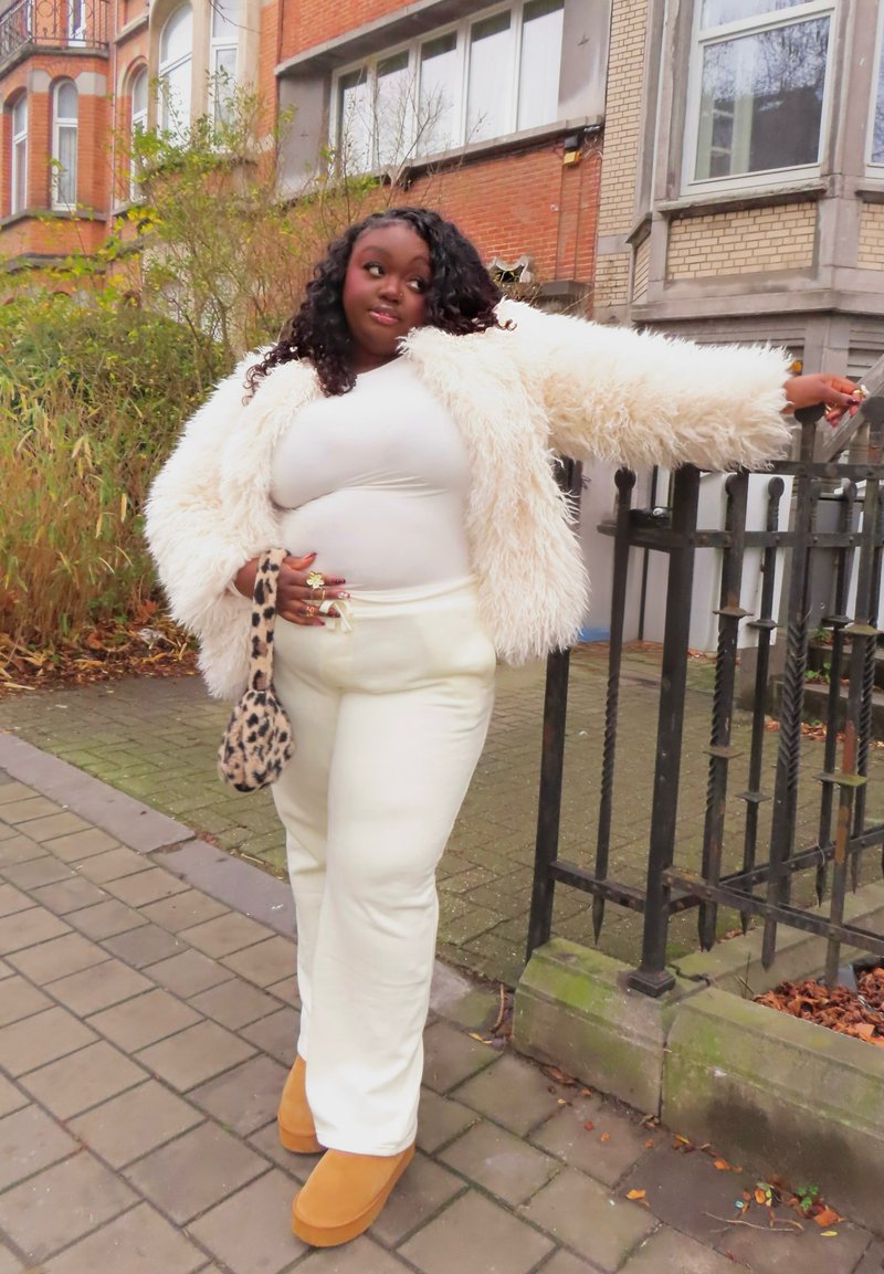 Femme aux cheveux bouclés portant une veste en peluche blanche, un haut blanc et un pantalon, appuyée contre une clôture en fer noir, tenant un sac à motif léopard sur un trottoir urbain.