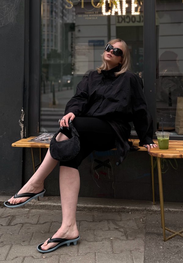 Vrouw in een oversized zwart overhemd, zonnebril en sandalen met hakken, zittend op een bankje buiten een café, houdt een zwarte tas vast met een groene drank in de buurt.