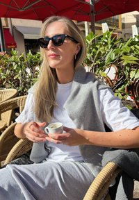 Vrouw zittend in een rotan stoel, met een klein wit koffie kopje in haar hand. Ze draagt een grijze trui over een wit T-shirt en heeft lang, steil haar.