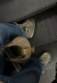 Sive kožne cipele s vezicama i gumanim potplatima, plave traperice i torba s uzorkom s bež remenom na pokretnim stepenicama.