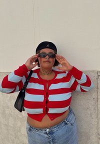 Cardigan rayé rouge et bleu clair avec des boutons, associé à un short en denim taille haute. Porte des lunettes de soleil noires et une casquette noire.