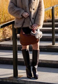 Femme portant un manteau en fourrure beige, une jupe en cuir noir, des bottes hautes noires, tenant un sac à main marron avec un pompon en fourrure près d'escaliers en béton en extérieur.