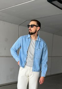 Chaqueta de mezclilla azul claro con cuello, un bolsillo en el pecho, usada sobre una camiseta gris. También se incluyen pantalones de color claro y gafas de sol negras.