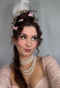 Collana di perle elegante a tre strati, abito off-the-shoulder rosa pallido con delicati accenti floreali, accessorio per capelli decorato con una piuma bianca.