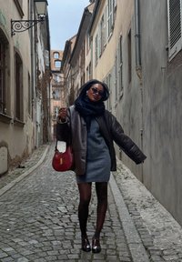 Hnědá kožená oversized bunda přes šedé šaty, černé punčocháče a lesklé černé boty. Drží červenou tašku a stojí na kostkované ulici.