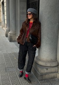 Femme appuyée contre une colonne en pierre, portant un béret gris, une veste marron à pois, un pull rouge, un pantalon noir ample, des chaussettes rouges et des mocassins noirs.