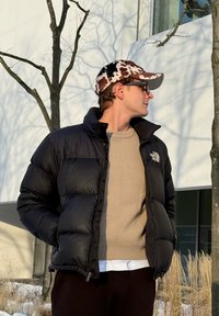 Homme portant une doudoune noire, un pull beige et une casquette à motif vache, debout à l'extérieur près d'arbres sans feuilles et d'herbes hautes.