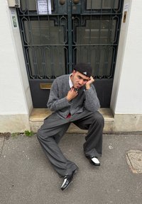 Uomo con cardigan grigio, pantaloni a righe sottili e basco nero seduto sul marciapiede davanti a un cancello di metallo, con la testa poggiata sulla mano e un'espressione seria.