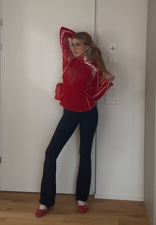 Una mujer con gafas posa en el interior vistiendo un suéter rojo, pantalones negros, zapatos rojos y lleva un bolso rojo al hombro.