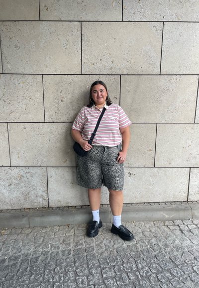 Camisa polo de manga corta a rayas en rosa y crema; pantalones cortos con estampado animal; zapatos negros; calcetines grises; de pie contra una pared de piedra.