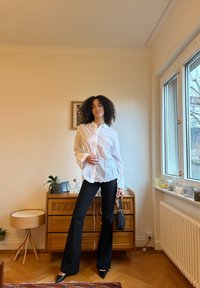 White button-up shirt with puffed sleeves, paired with black flared trousers and pointed black heels. Small black handbag in hand.