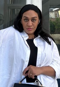 Hvit oversized skjorte over en svart topp, med en teksturert svart håndveske holdt i den ene hånden. Håret er mørkt og bølgete, komplettert med hoopsøljer.