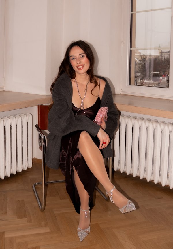 Joven mujer con vestido oscuro, cárdigan gris y tacones brillantes sentada en una silla junto a la ventana sosteniendo un bolso rosa, sonriendo a la cámara.