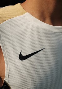 Hvit mesh idrettstopp med en teksturert overflate og svart Nike swoosh-logo; har en beige detalj på skulderen.