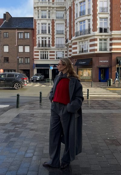 Mujer con abrigo gris y suéter rojo de pie en la acera de la ciudad cerca de un paso de peatones, con edificios históricos y coches de fondo.