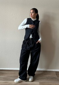 Donna con capelli scuri raccolti in una coda di cavallo indossa un gilet senza maniche nero sopra una maglietta bianca a maniche lunghe, pantaloni neri e sneakers bianche, in piedi contro un muro beige.