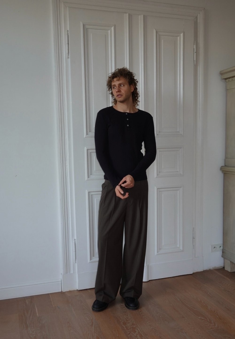 Camicia nera a maniche lunghe con bottoni, abbinata a pantaloni larghi marroni a righe sottili e scarpe nere. Pavimenti in legno e porte bianche a pannelli sullo sfondo.